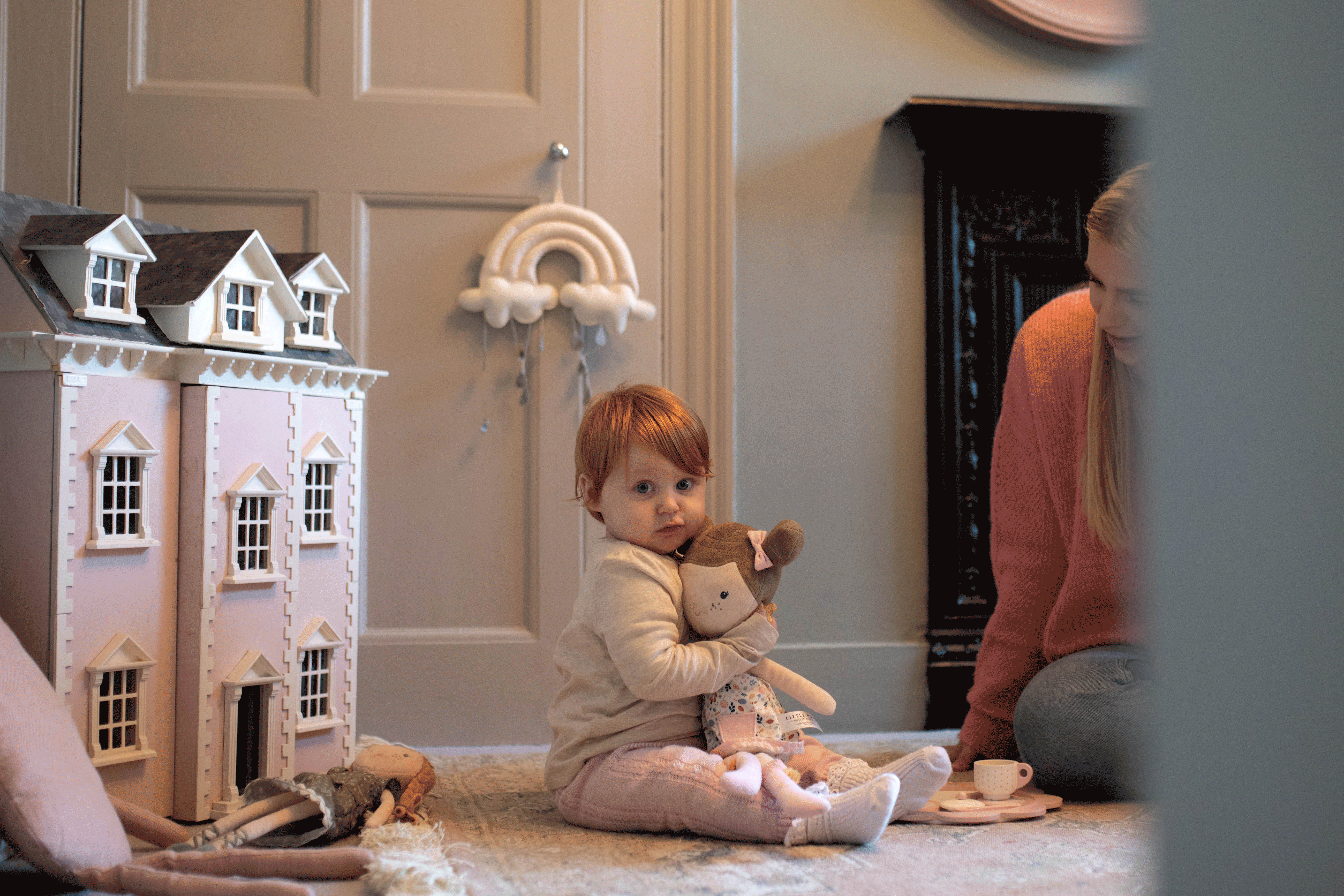 A toddler sitting on the rug holding a rag doll next to a detailed pink wooden dollshouse, with an adult watching nearby.