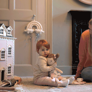 A toddler sitting on the rug holding a rag doll next to a detailed pink wooden dollshouse, with an adult watching nearby.
