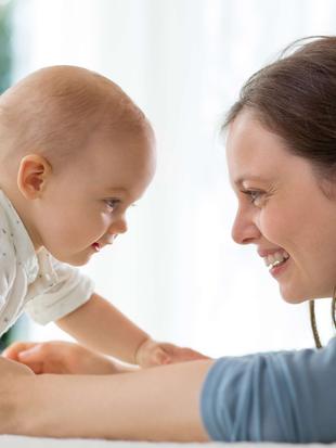 Ein zärtlicher Moment zwischen einer Mutter und ihrem Baby, in dem das Baby die Hand ausstreckt, um das Gesicht der Mutter zu berühren, und die Mutter zärtlich lächelt.