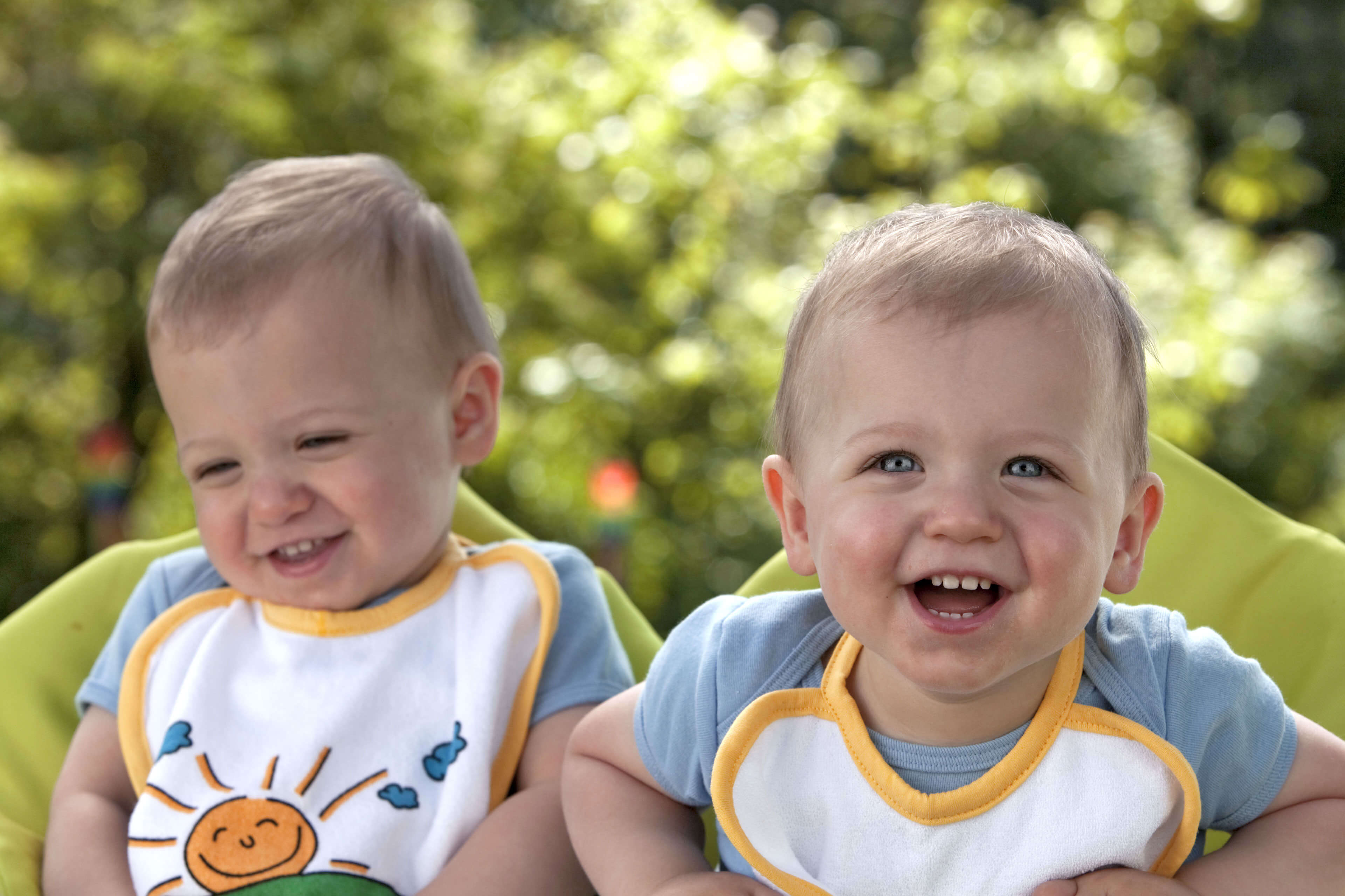 Zwei Kleinkinder, eines lachend und das andere lächelnd, im Freien mit Grün im Hintergrund.