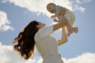 Mutter hebt ihr lachendes Baby im Freien in die Luft, beide genießen einen unbeschwerten Moment unter blauem Himmel