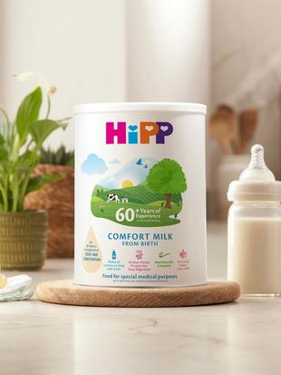 HiPP Comfort milk tub on a bright kitchen worktop beside a prepared baby bottle, dummy, muslin cloth, plant and soft toy