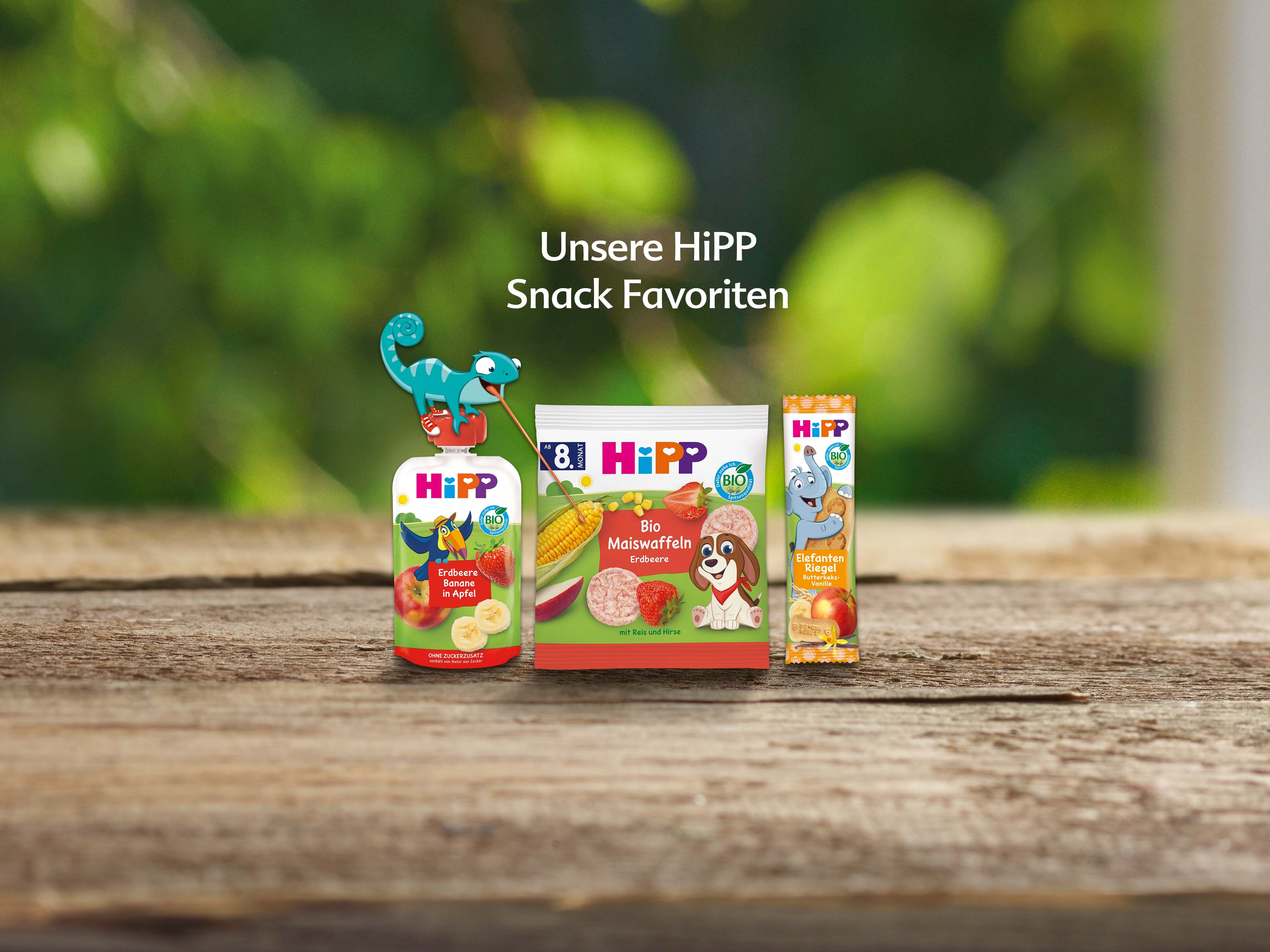 Drei HiPP-Snackprodukte auf Holzuntergrund vor unscharf grünem Hintergrund mit Text 'Unsere HiPP Snack Favoriten'