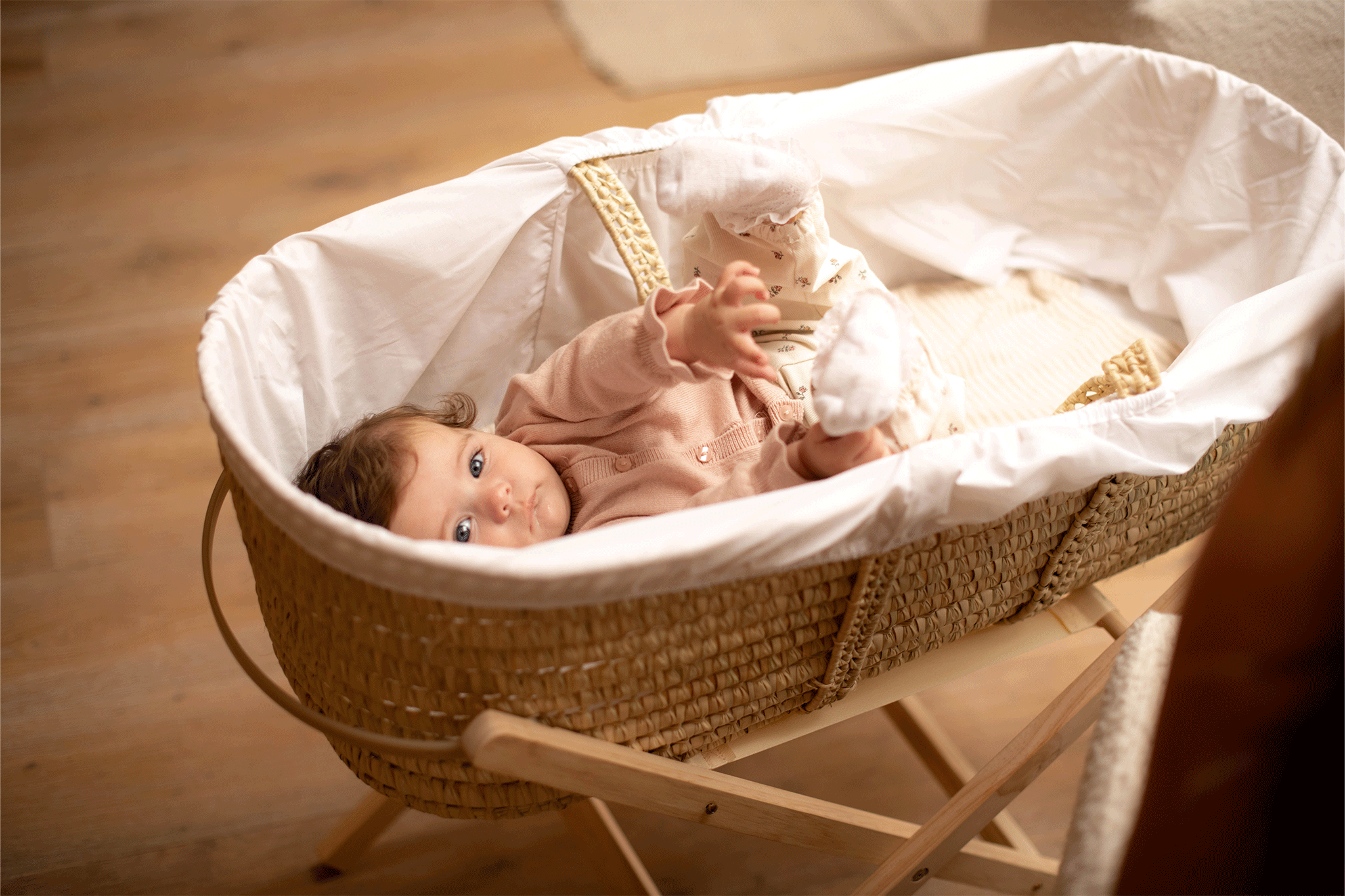 A baby wearing a light pink cardigan lying on back inside a woven Moses basket resting on a wooden stand.