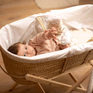 A baby wearing a light pink cardigan lying on back inside a woven Moses basket resting on a wooden stand.
