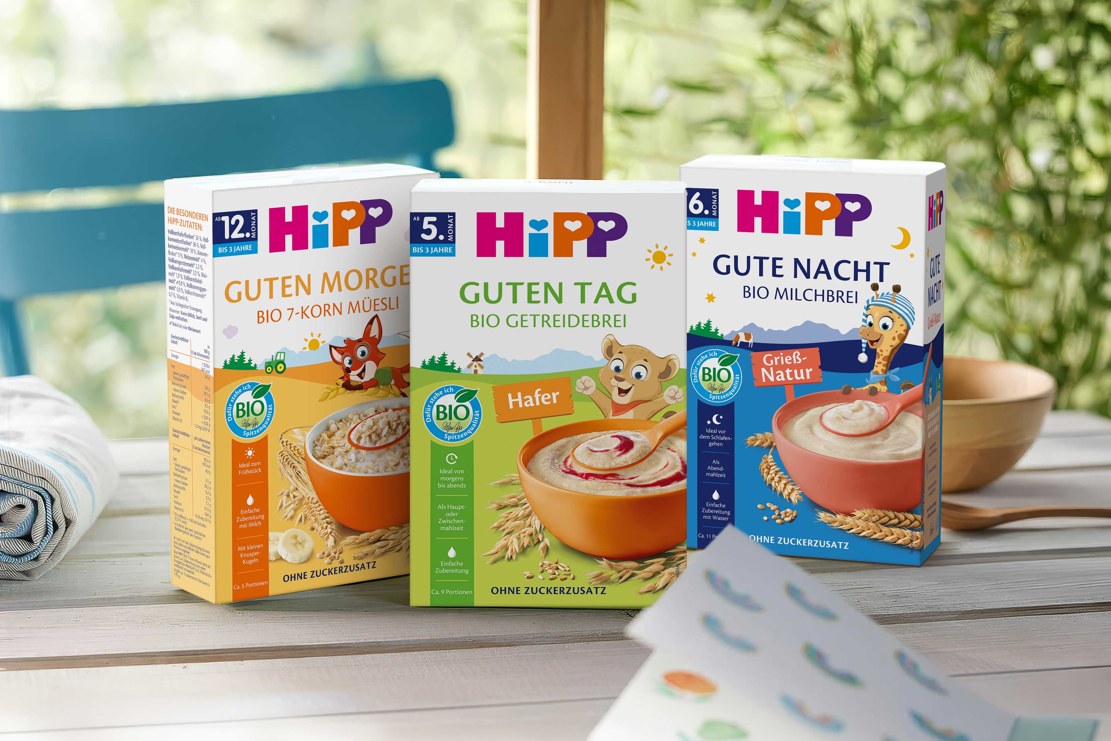 Drei Packungen HiPP Bio-Brei mit den Aufschriften 'Guten Morgen', 'Guten Tag' und 'Gute Nacht' stehend auf einem Holztisch vor einem Fenster