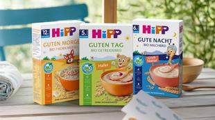 Drei Packungen HiPP Bio-Brei mit den Aufschriften 'Guten Morgen', 'Guten Tag' und 'Gute Nacht' stehend auf einem Holztisch vor einem Fenster