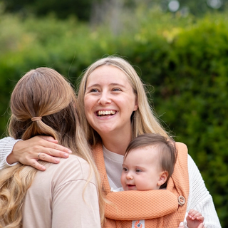 Smiling parent wearing a baby in a soft carrier, hugging a friend outdoors, sharing a warm, supportive moment.