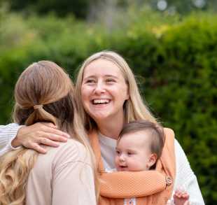 Smiling mum cuddling a friend while carrying her baby in a soft orange carrier outdoors