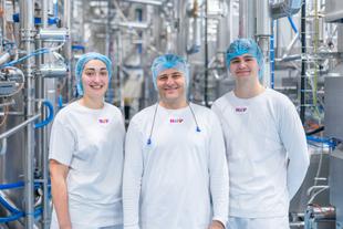 Drei Personen in weißen T-Shirts mit buntem HiPP-Logo und blauen Haarnetzen stehen in einer Produktionsanlage mit vielen Rohren und Maschinen