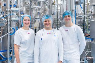 Drei Personen in weißen T-Shirts mit buntem HiPP-Logo und blauen Haarnetzen stehen in einer Produktionsanlage mit vielen Rohren und Maschinen