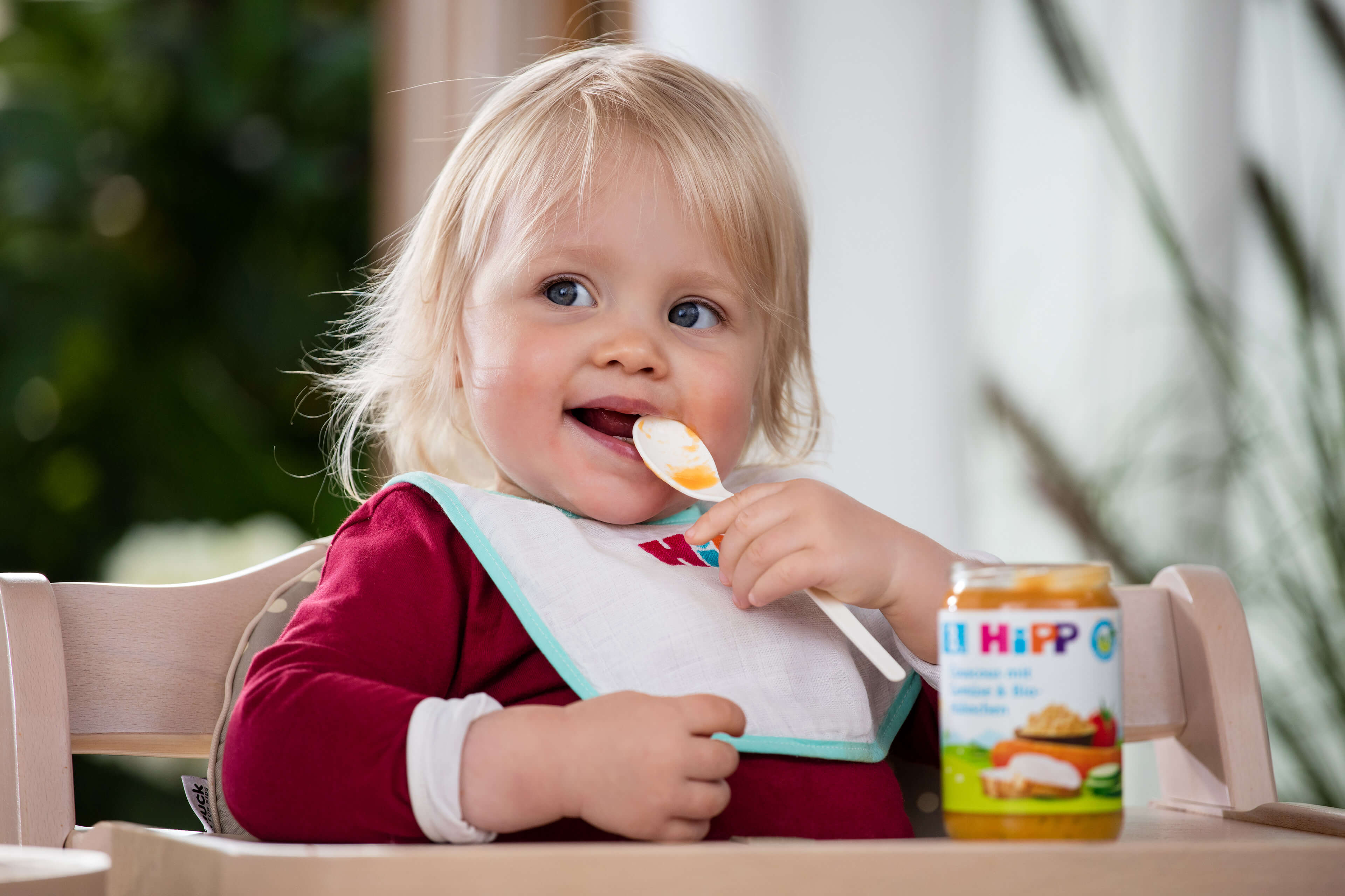Kleines Kind mit Lätzchen sitzt am Tisch und hält einen Löffel neben einem Hipp-Babynahrungsglas.