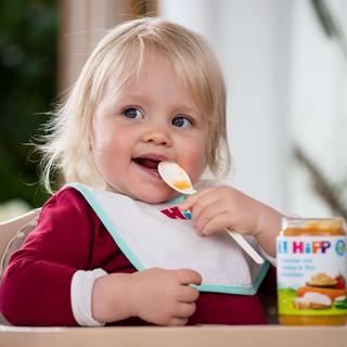 Kind sitzt auf einem Hochstuhl und hält ein Lätzchen mit einer Hand, auf dem Tisch steht ein Glas Babynahrung der Marke HiPP