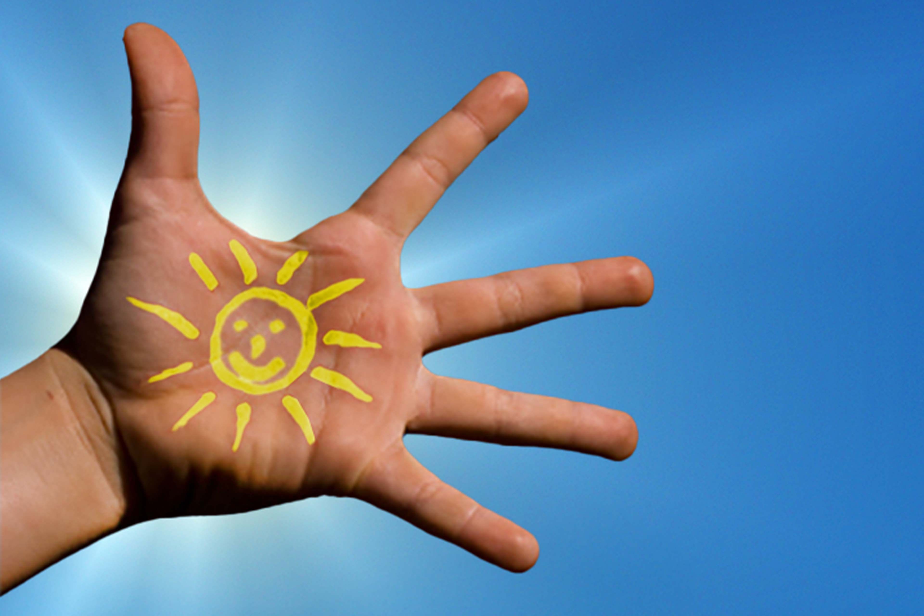 Eine Hand mit einer aufgemalten Sonne vor einem strahlend blauen Himmel.