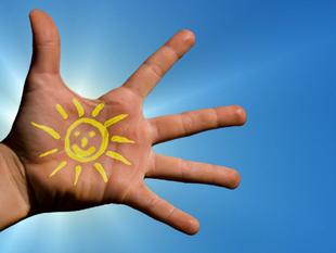 Eine Hand mit einer aufgemalten Sonne vor einem strahlend blauen Himmel.