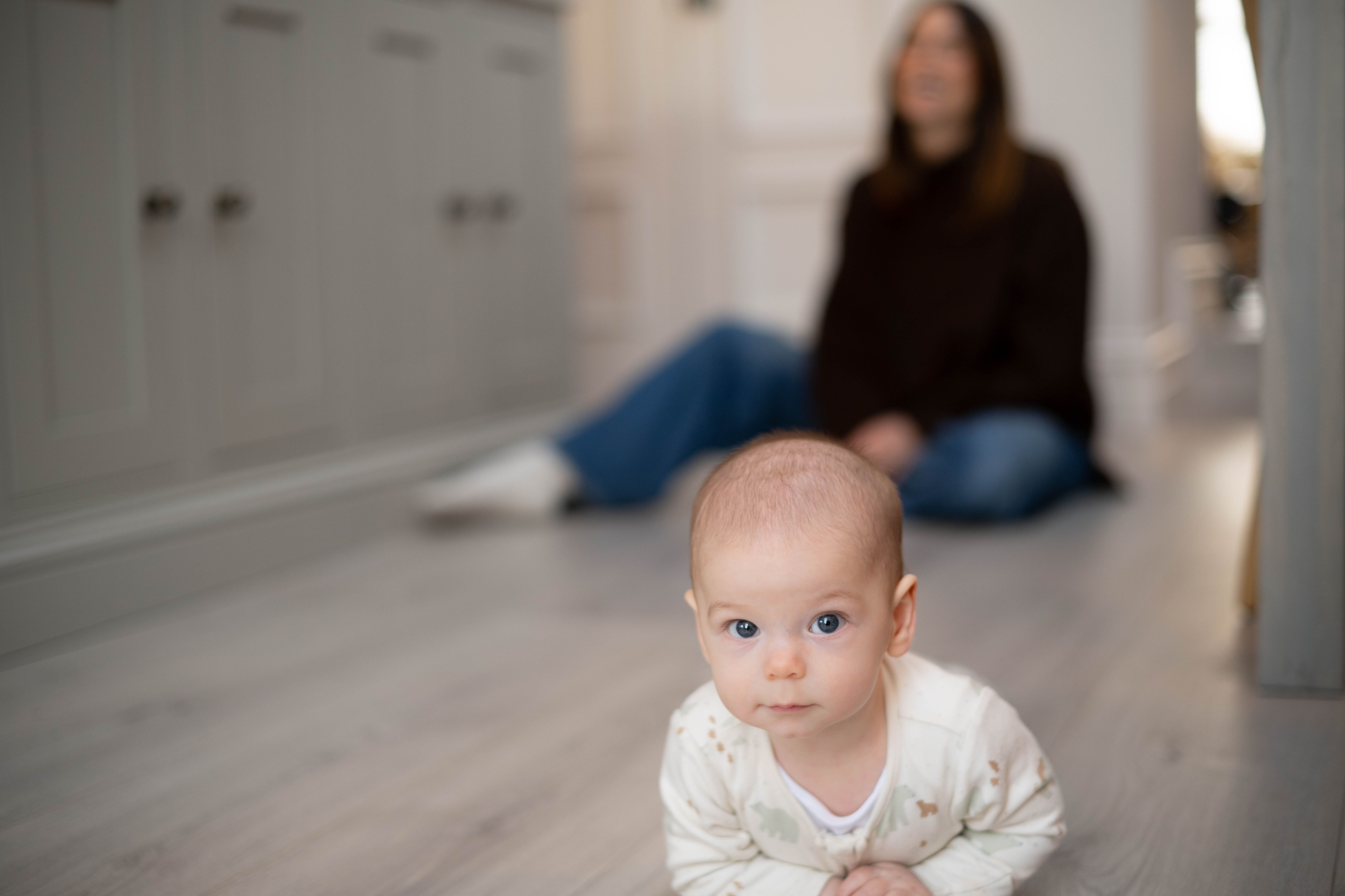 Young baby lying on their tummy on a kitchen floor, watched closely by a seated parent in the background