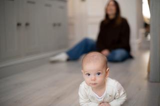 Young baby crawling/on their tummy looking at the camera with mum in background