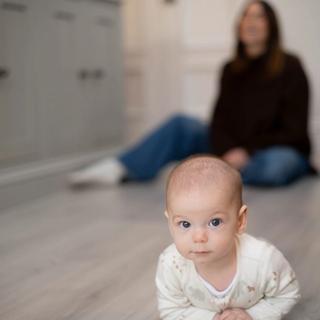 Young baby lying on their tummy on a kitchen floor, watched closely by a seated parent in the background
