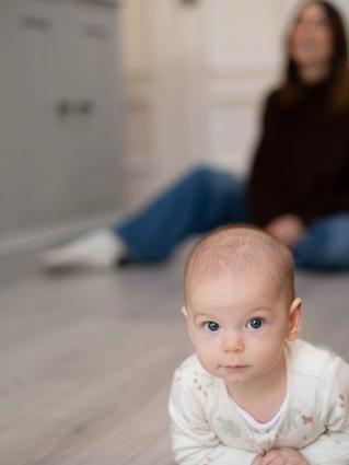 Young baby lying on their tummy on a kitchen floor, watched closely by a seated parent in the background