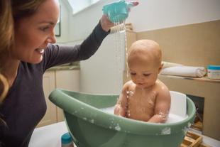 Frau gießt Wasser aus einem Becher über ein Baby, das in einer Babybadewanne sitzt