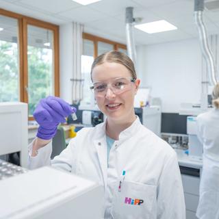 Lächelnde HiPP-Chemielaborantin im weißen Kittel hält im Labor ein Probenröhrchen hoch und arbeitet an moderner Analytiktechnik.