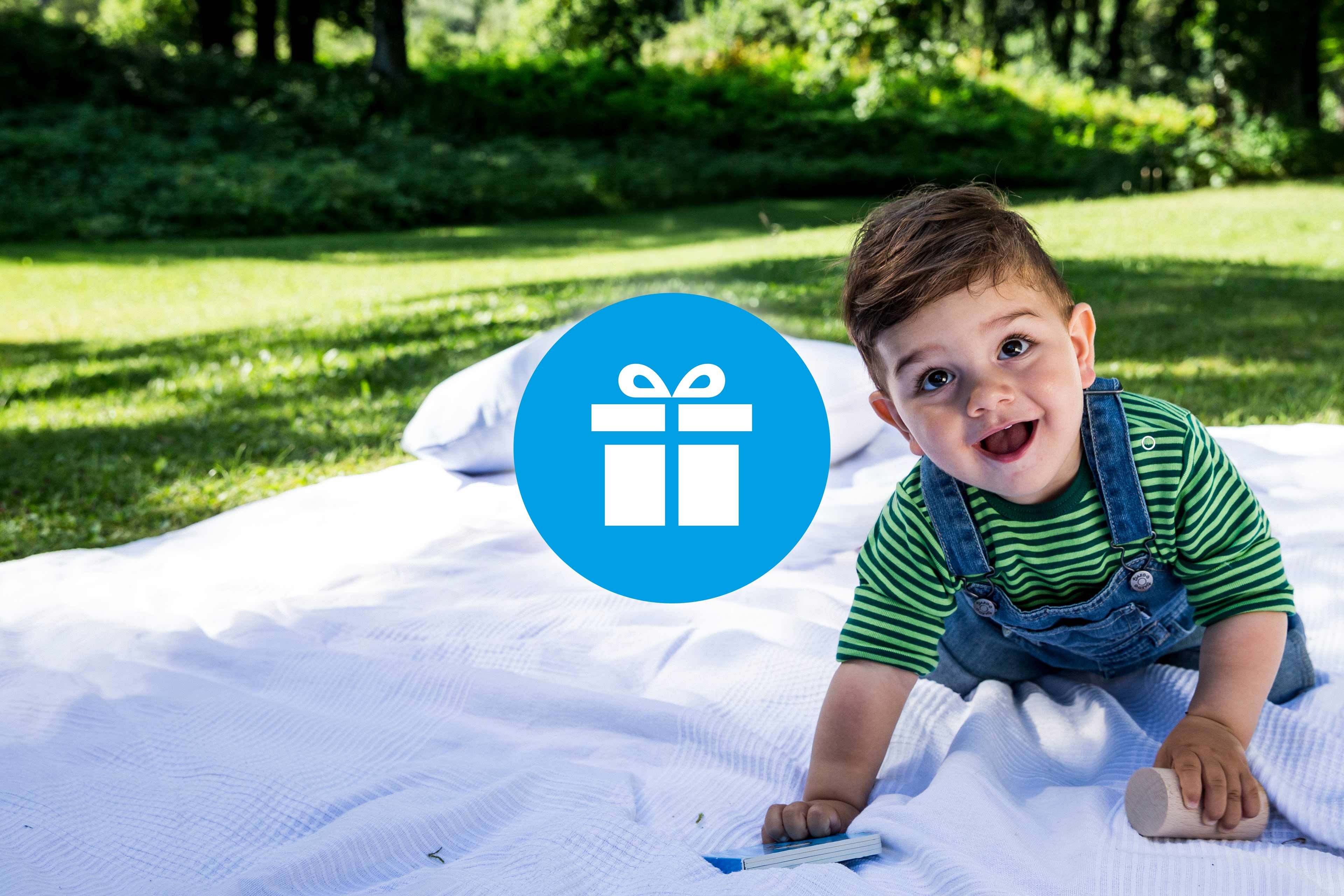 Lachendes Baby auf einer Picknickdecke im Grünen, daneben ein Geschenk-Symbol als Hinweis auf Gewinnspiele