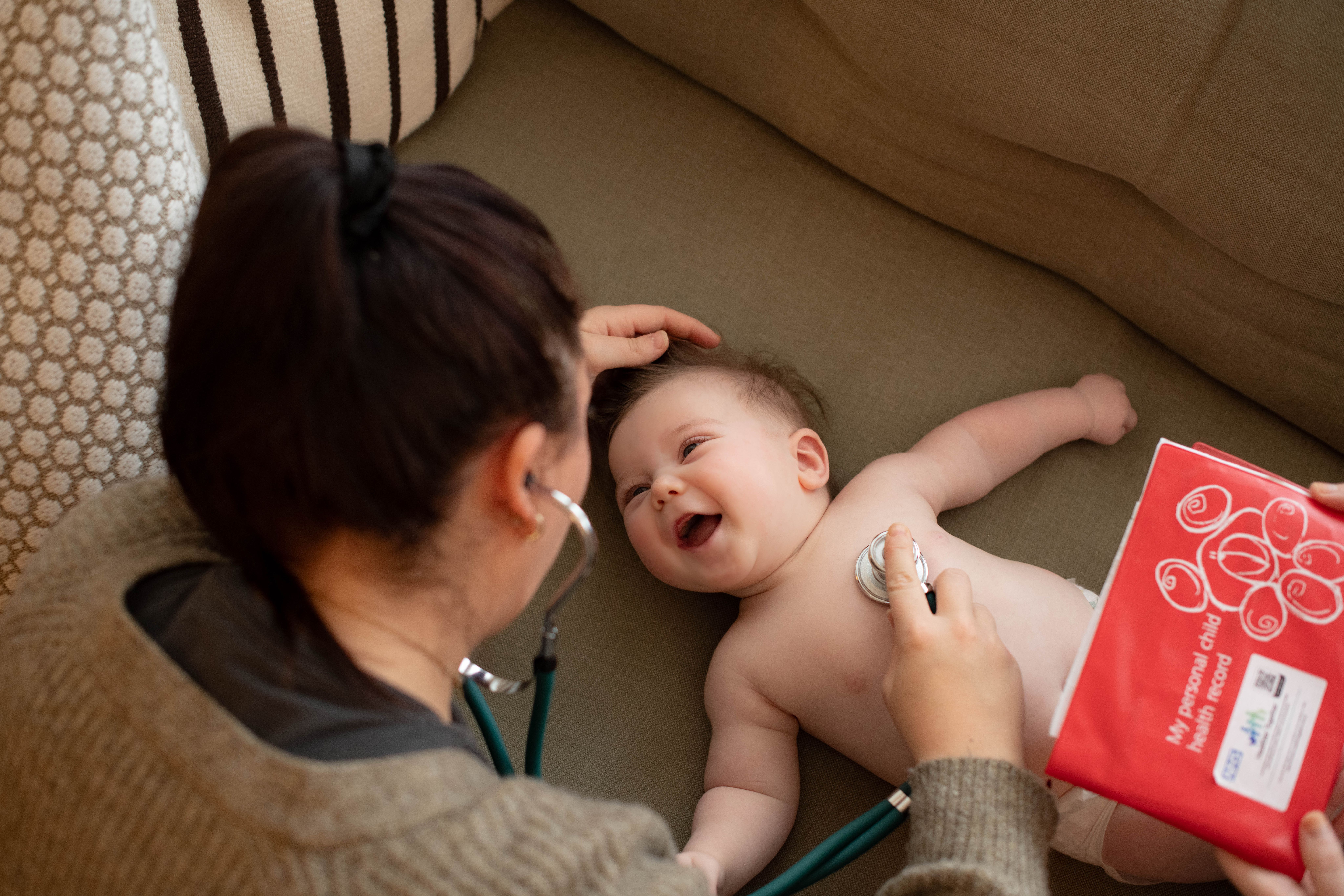 Smiling baby lying on a sofa while a healthcare professional listens to their chest with a stethoscope and holds health notes.