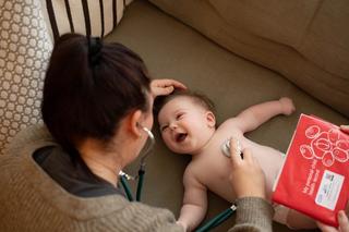 Healthcare professional with a stethoscope listening to a smiling baby's chest, with the red health visitor book on show
