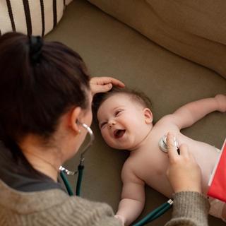Smiling baby lying on a sofa while a healthcare professional listens to their chest with a stethoscope and holds health notes.