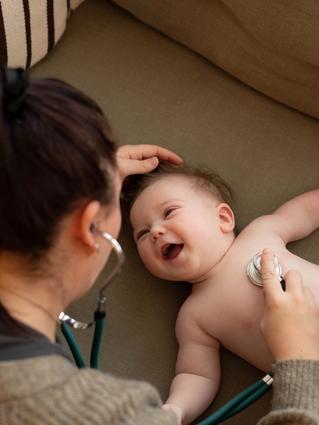 Smiling baby lying on a sofa while a healthcare professional listens to their chest with a stethoscope and holds health notes.