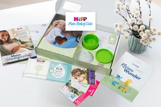 Geöffnete Geschenkbox mit Babyprodukten, Feuchttüchern, Broschüren und einem Buch neben einem Zweig mit Weidenkätzchen auf weißem Untergrund