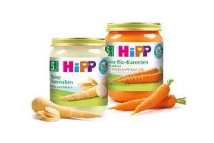 Zwei Gläschen Babynahrung mit der Aufschrift "Hipp" mit verschiedenen Obst- und Gemüsesorten, dazu frische Karotten.
