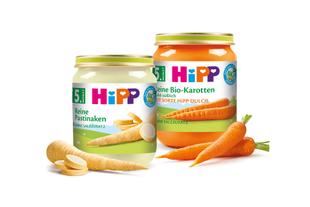 Zwei Gläschen Babynahrung mit der Aufschrift "Hipp" mit verschiedenen Obst- und Gemüsesorten, dazu frische Karotten.