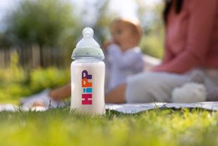 Babyflasche mit der Aufschrift 'HiPP' steht auf Gras, im Hintergrund unscharf sitzende Frau und Kleinkind