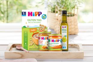 Vier verschiedene Hipp-Babynahrungsprodukte auf einem Holztablett vor unscharfem Hintergrund