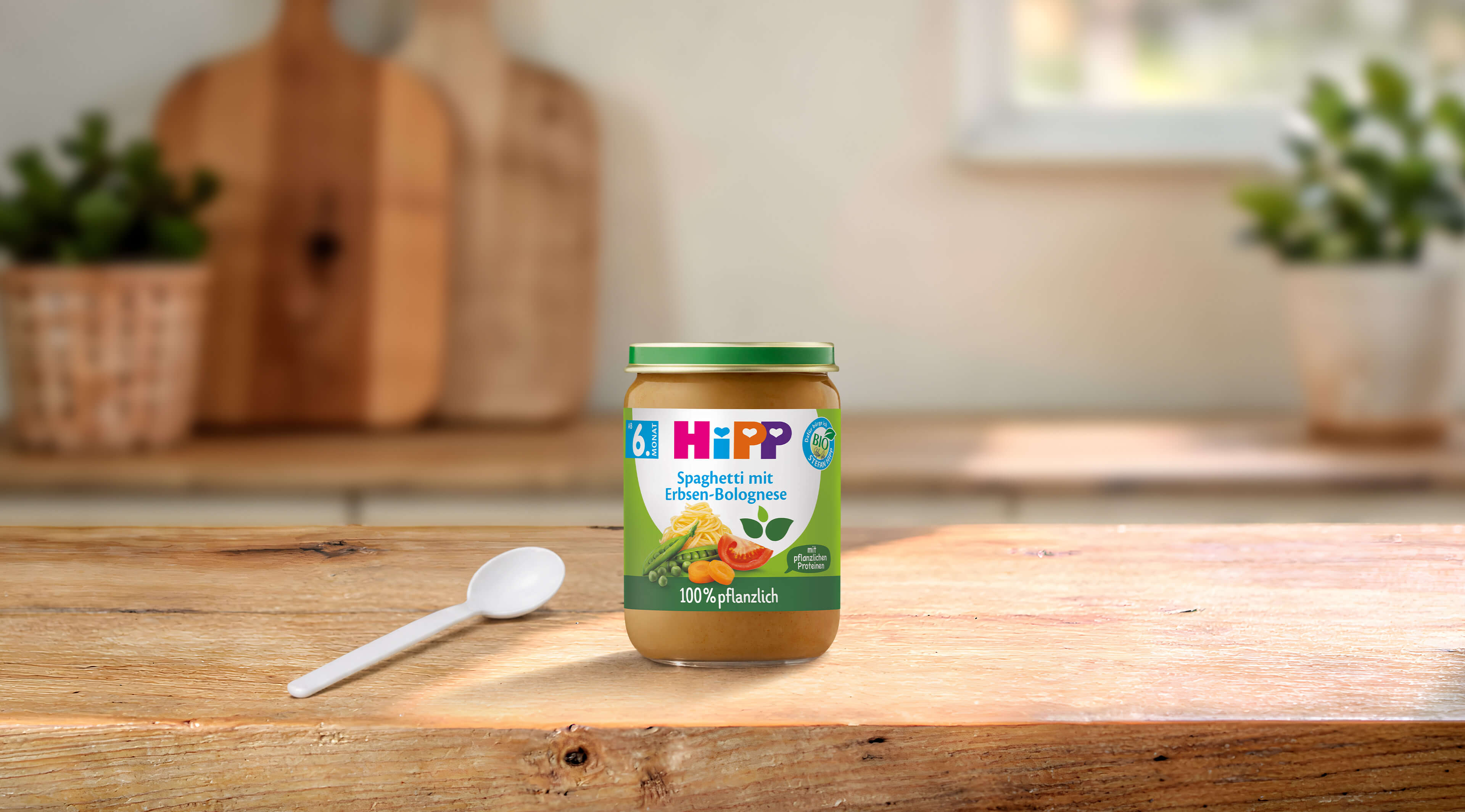 HiPP Bio-Gläschen Spaghetti mit Erbsen-Bolognese ab dem 6. Monat auf Holztisch mit Löffel in heller Küche