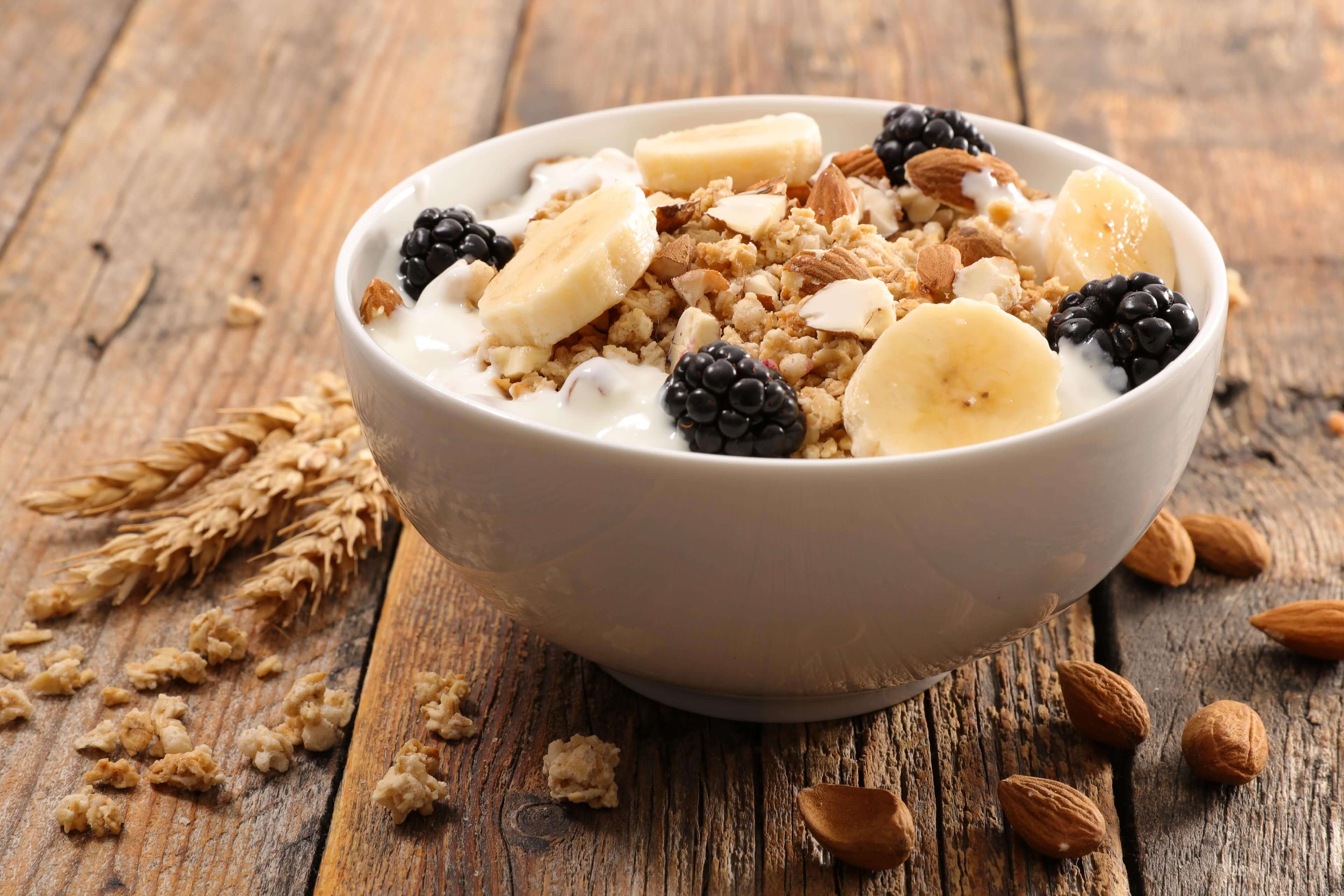 Eine Schale Frühstücksflocken mit Bananenscheiben, Brombeeren und Mandeln, begleitet von einem Weizenstängel und Mandeln auf einer rustikalen Holzunterlage.