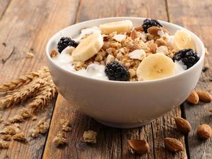 Eine Schale Frühstücksflocken mit Bananenscheiben, Brombeeren und Mandeln, begleitet von einem Weizenstängel und Mandeln auf einer rustikalen Holzunterlage.
