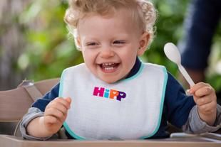 Ein fröhliches Kleinkind, das einen Löffel hält und ein Lätzchen mit den Buchstaben "HIPP" trägt.