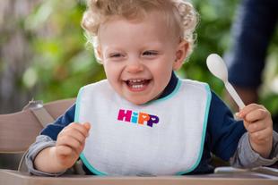 Ein fröhliches Kleinkind, das einen Löffel hält und ein Lätzchen mit den Buchstaben "HIPP" trägt.