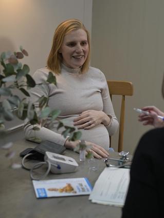 A smiling pregnant woman sits at a table during a prenatal appointment, resting a hand on her belly while chatting with a healthcare professional. A blood pressure cuff, stethoscope, and medical forms are on the table.