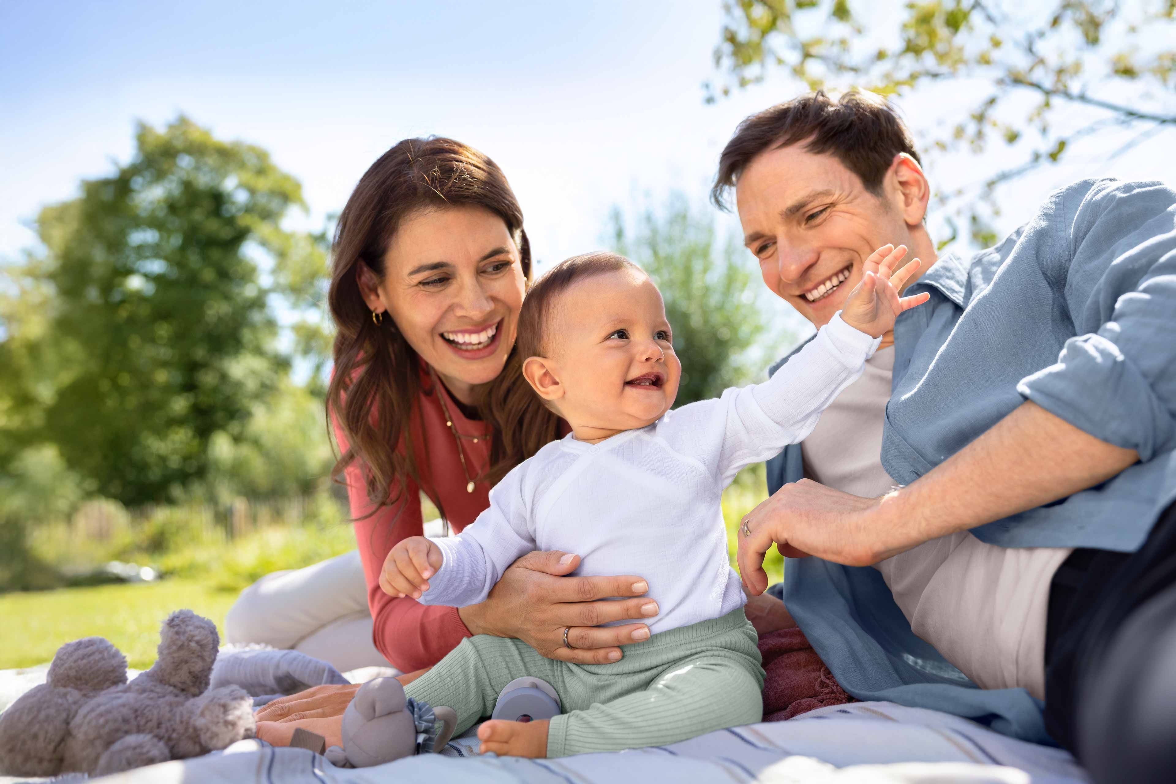 Ein freudiger Familienmoment im Freien mit einer Mutter und einem Vater, die ihr Baby anlächeln, das sich ihnen entgegenstreckt.