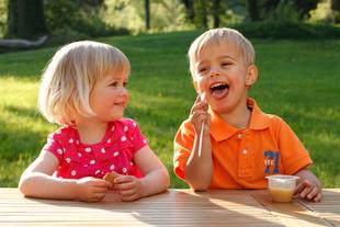 Zwei kleine Kinder, ein Mädchen und ein Junge, sitzen im Freien an einem Holztisch, wobei der Junge etwas isst und das Mädchen ein kleines Gefäß hält.