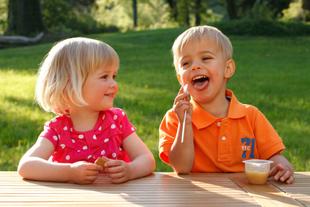 Zwei kleine Kinder, ein Mädchen und ein Junge, sitzen im Freien an einem Holztisch, wobei der Junge etwas isst und das Mädchen ein kleines Gefäß hält.