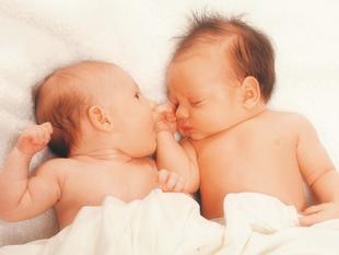 Zwei Säuglinge liegen dicht nebeneinander auf einer weißen Fläche, wobei ein Baby die Hand des anderen berührt.