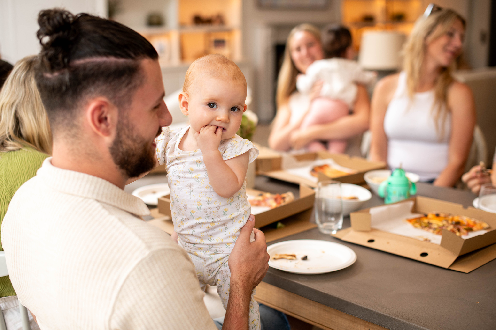 A man holding a baby in patterned babygrows, who is sucking on their fingers while sitting at a dining table surrounded by family and open takeaway pizza boxes.