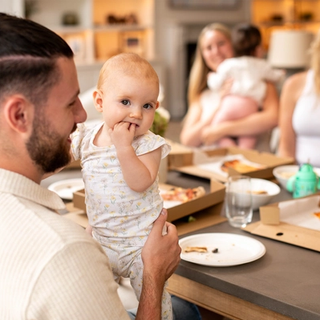 A man holding a baby in patterned babygrows, who is sucking on their fingers while sitting at a dining table surrounded by family and open takeaway pizza boxes.