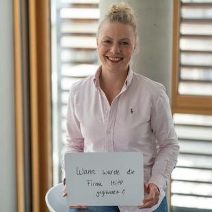 HiPP Mitarbeiterin hält ein Schild mit der handschriftlichen Frage 'Wann wurde die Firma Hipp gegründet'