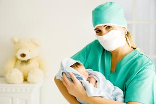 Eine medizinische Fachkraft in grüner Kleidung hält ein neugeborenes Baby, mit einem Teddybär im Hintergrund.