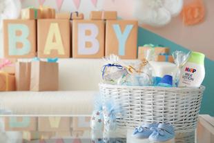 Ein Babyparty-Setup mit einem weißen Weidenkorb, gefüllt mit Babyartikeln, einschließlich einer Flasche Milchlotion, Babyschuhen und anderen wichtigen Babyartikeln, die auf einer reflektierenden Oberfläche platziert werden.